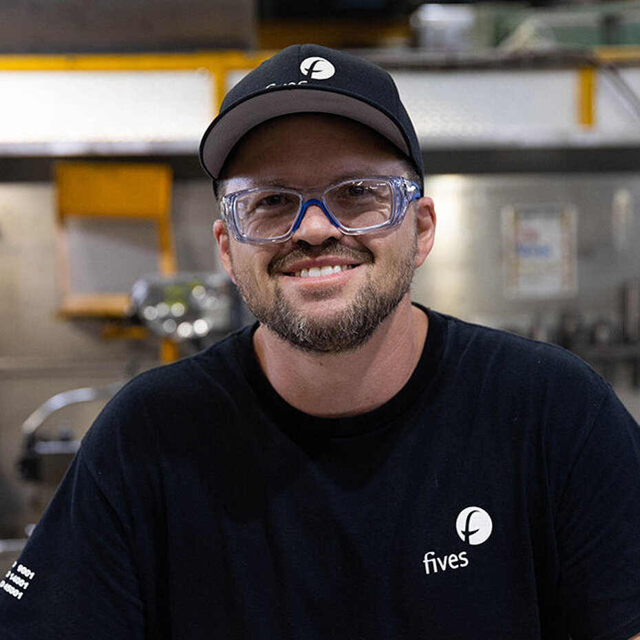 Portrait d’un employé portant des lunettes de sécurité et une casquette noire Fives, souriant dans un atelier industriel.