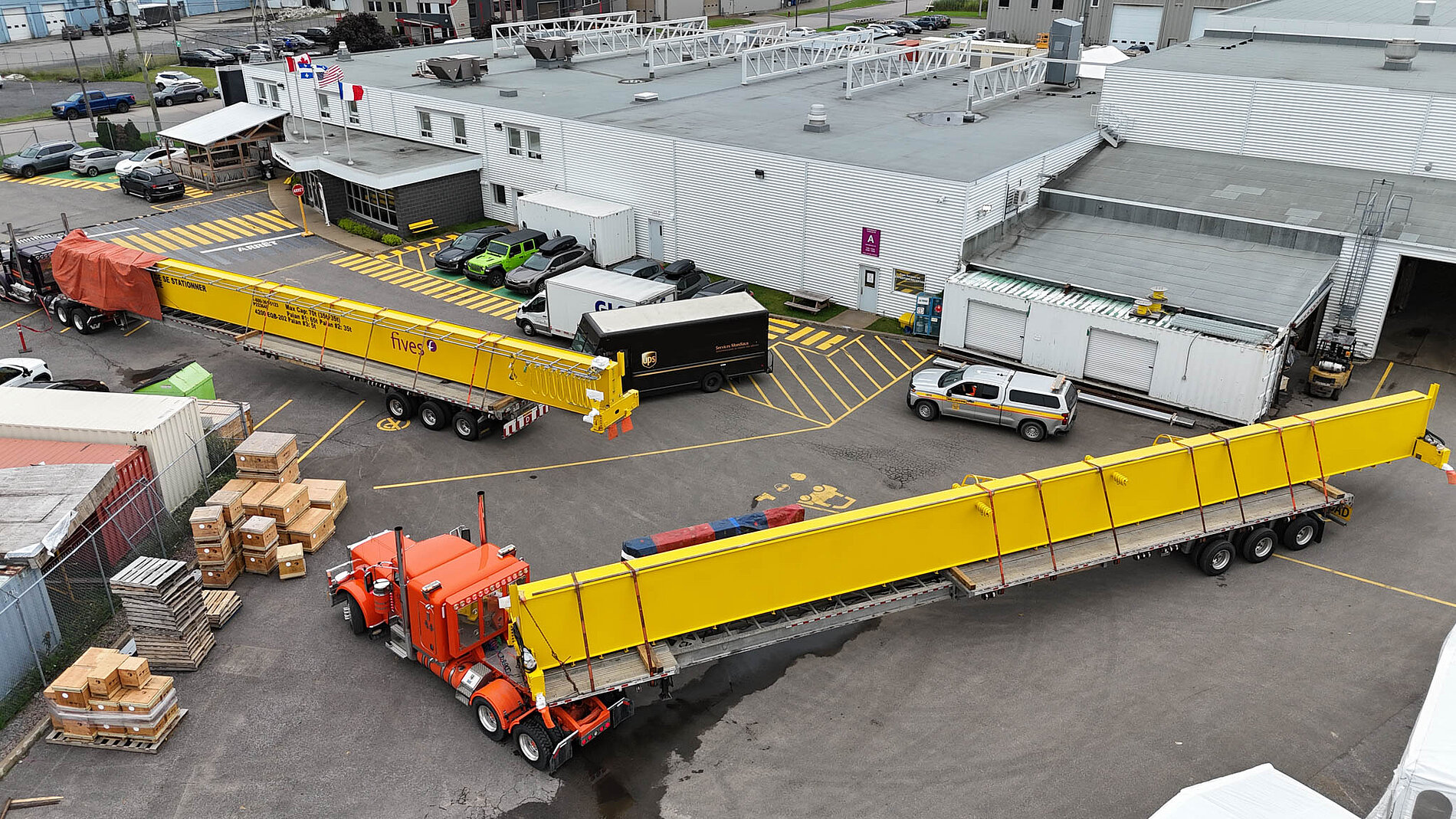 Aerial view of two large yellow overhead crane girders leaving an industrial workshop on orange transport trucks, surrounded by warehouses, shipping containers, and stacked materials in a manufacturing yard.